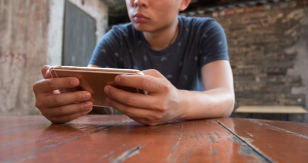 Ein Junge hat seine Hände auf einem Holztisch aufgestützt und hält dabei ein iPhone in seinen Händen.
